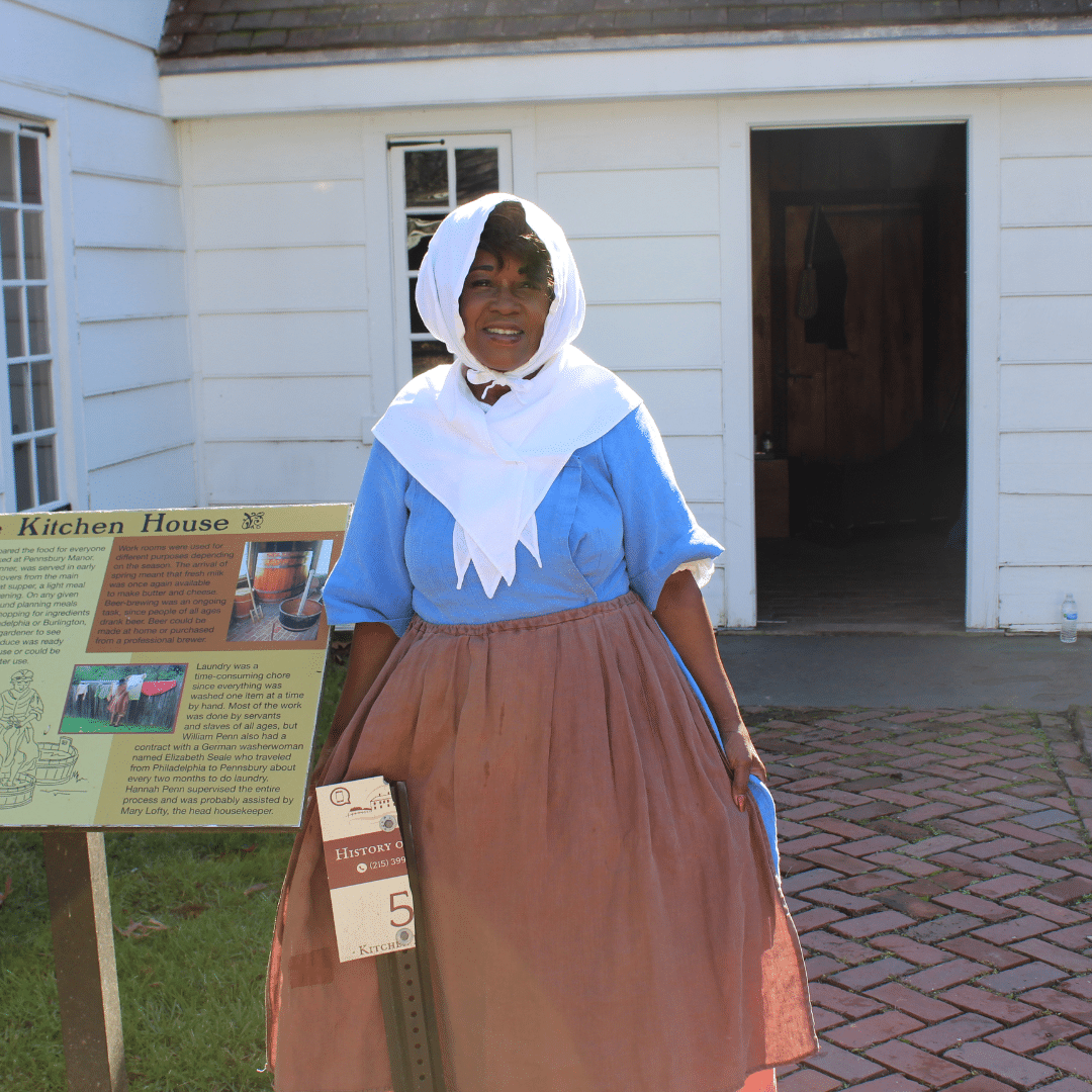 A woman dressed in colonial attire stands outside the kitchen house