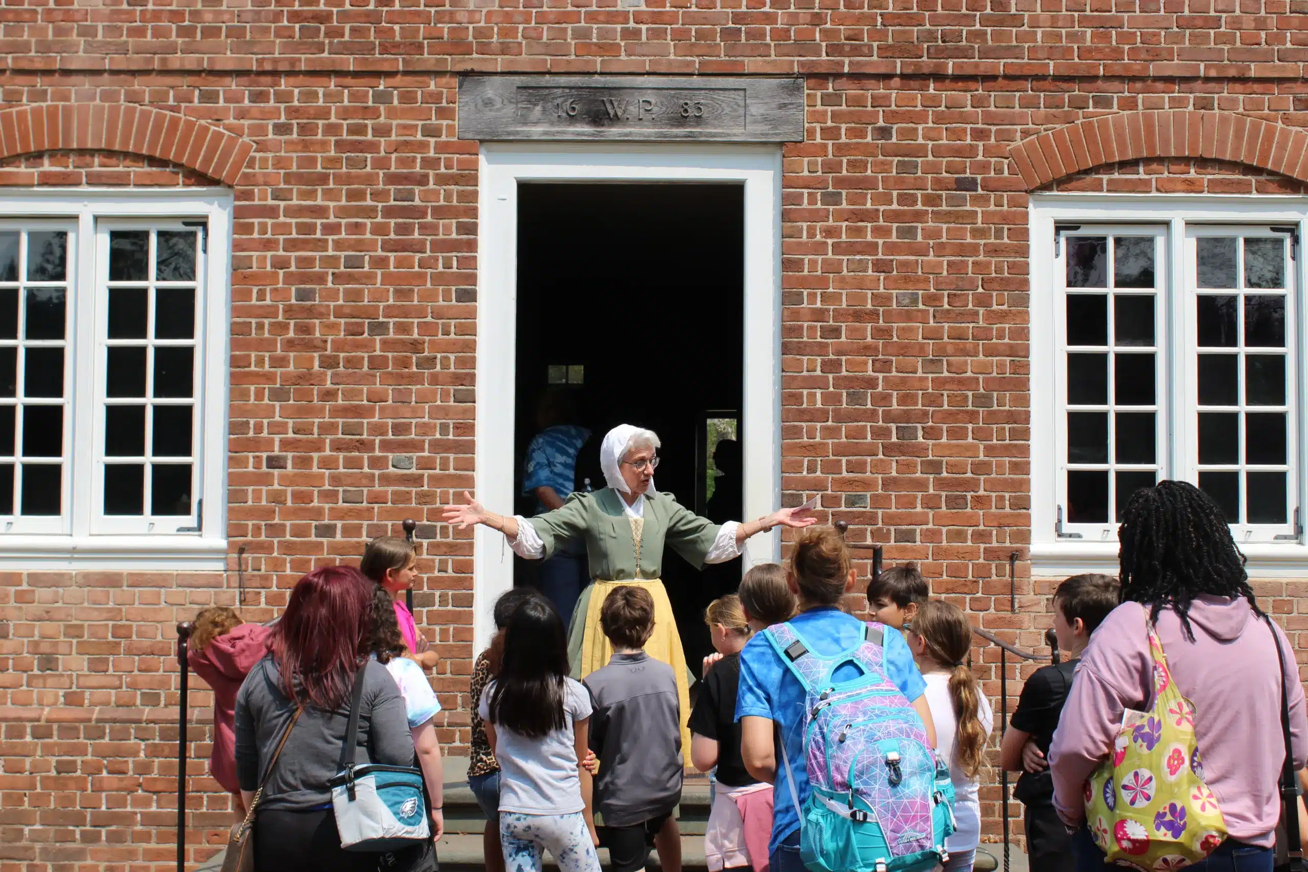 A woman dressed in colonial attire stands outside the manor house entrance with her arms open to welcome a group of students to the Manor House.