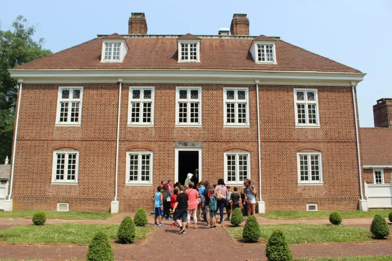 A group of kids on a school field trip stands outside William Penn's Manor house with a tour guide at the entrance.